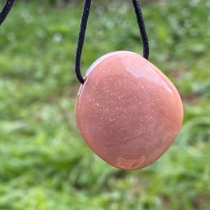 Un pendentif en pierre polie, de forme galet, suspendu à un cordon noir. La pierre est de couleur pêche-rosé avec des inclusions pailletées scintillantes, se détachant sur un arrière-plan flou de pelouse verte avec de petites fleurs blanches.