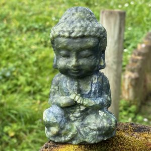 Statue de Bouddha sculptée en jade vert naturel, assis en position de méditation sur un morceau de bois dans un jardin zen.