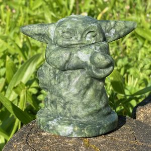 Photographie en gros plan d'une figurine sculptée de Grogu (Baby Yoda) en jade vert véritable, montrant la texture polie de la pierre, ses grandes oreilles et son expression paisible, sur un fond flou de nature lumineux.