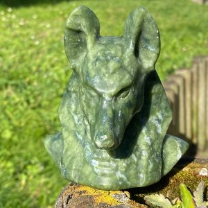 Sculpture réaliste d'une tête d'Anubis en jade vert sculpté, vue de face sur un fond flou de la nature verte.