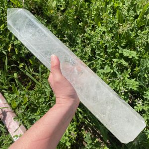 Grande tour polie en cristal de roche naturel transparent, forme obélisque hexagonal, posée sur une tranche de bois dans un jardin ensoleillé avec des fleurs.