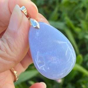 Pendentif en calcédoine bleue naturelle polie. La pierre présente une teinte bleu ciel laiteux avec de légères bandes translucides, suspendue à une bélière argentée sur un fond neutre et lumineux.